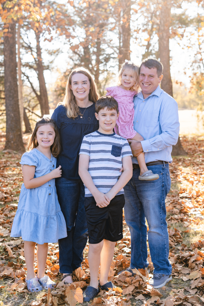 A family of five poses together outdoors in a park during autumn. Two adults stand with three children among fallen leaves and tall trees with golden-brown foliage.