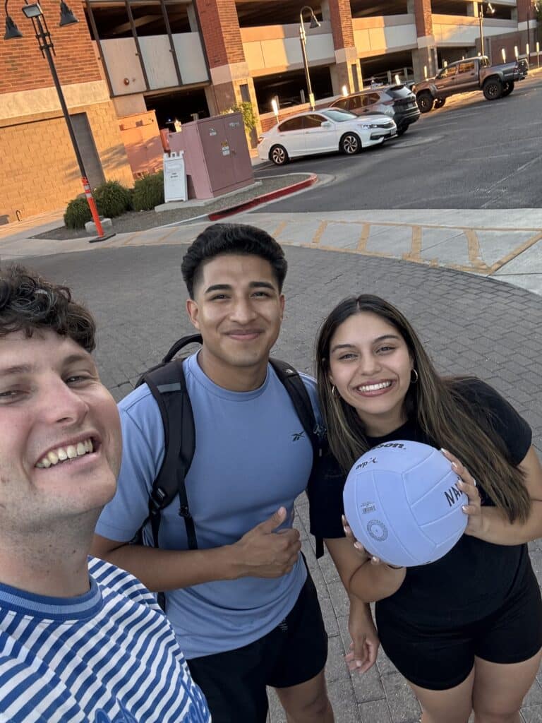Three friends pose for a selfie outside a parking garage, one holding a volleyball and all smiling at the camera.