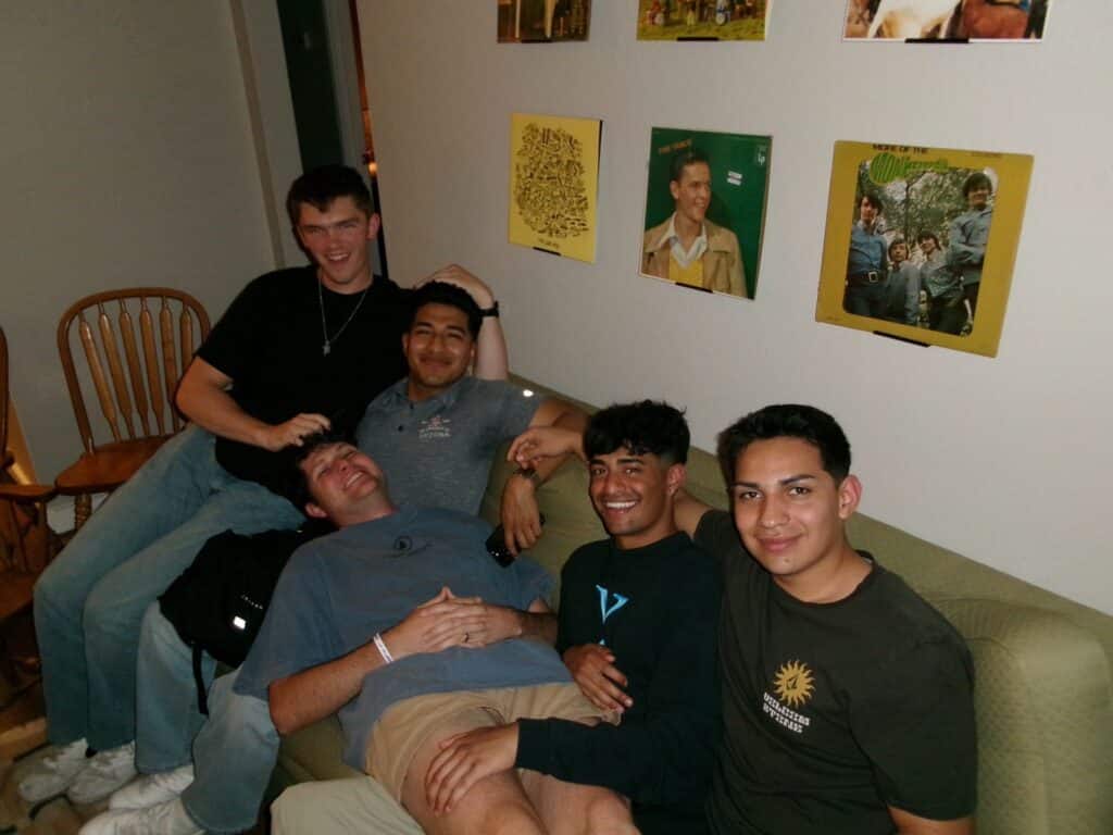 Five young men relax together on a couch in a living room, smiling at the camera.