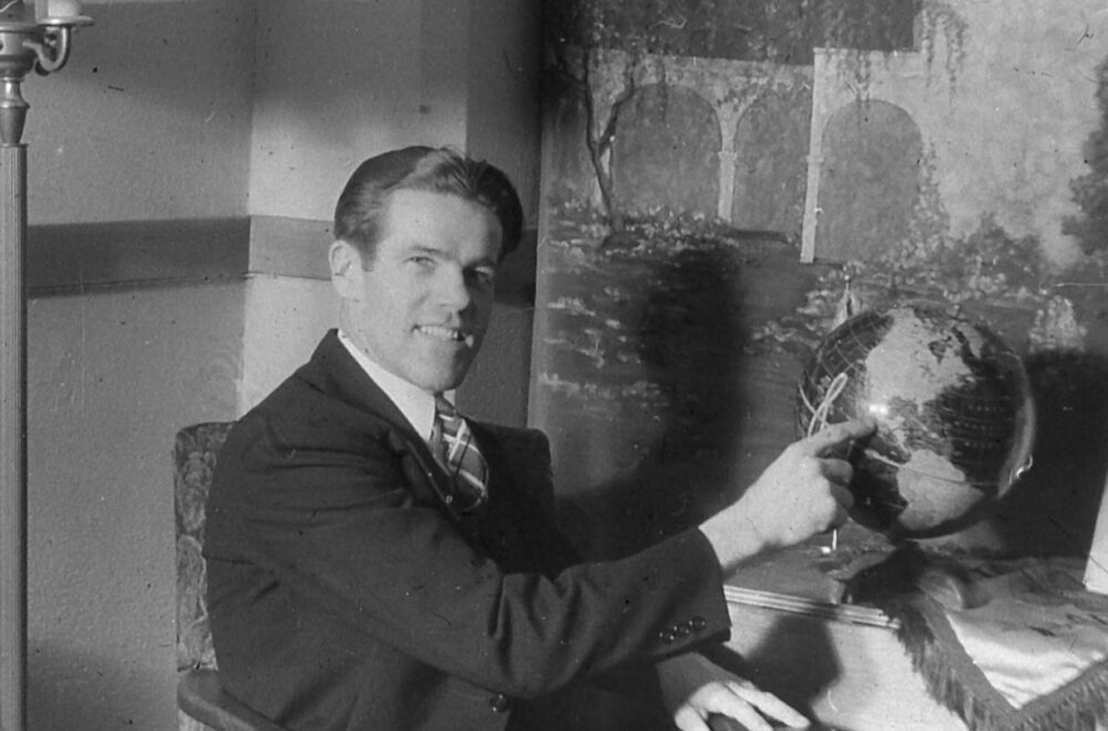Man in a suit sits at a desk, pointing to a globe in a vintage black-and-white photo.