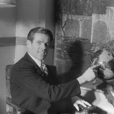Man in a suit sits at a desk, pointing to a globe in a vintage black-and-white photo.