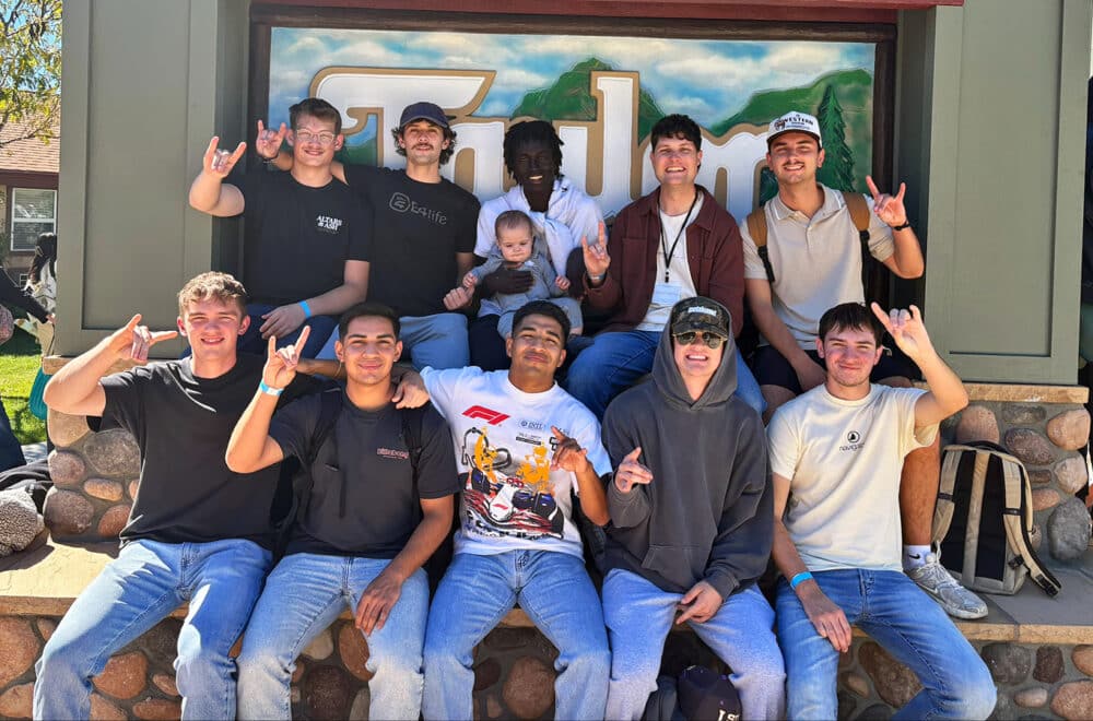 A group of 10 young men and one baby pose together in front of a rustic camp sign, smiling and making hand gestures.