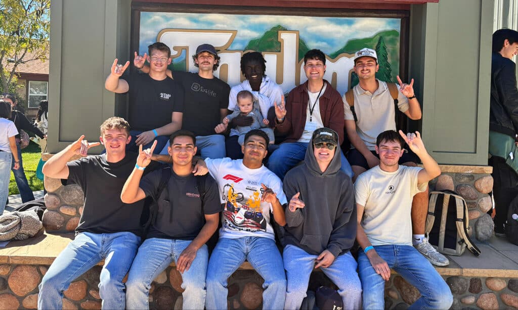 A group of 10 young men and one baby pose together in front of a rustic camp sign, smiling and making hand gestures.