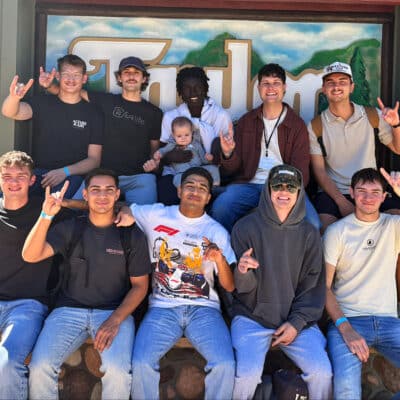 A group of 10 young men and one baby pose together in front of a rustic camp sign, smiling and making hand gestures.