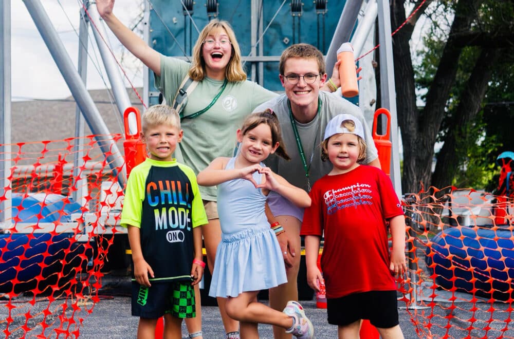 Group of smiling children with two camp counselors posing in front of a climbing structure at an outdoor camp, orange safety net behind them.
