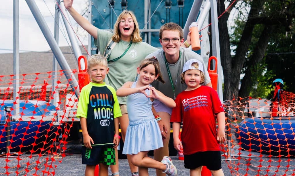 Group of smiling children with two camp counselors posing in front of a climbing structure at an outdoor camp, orange safety net behind them.