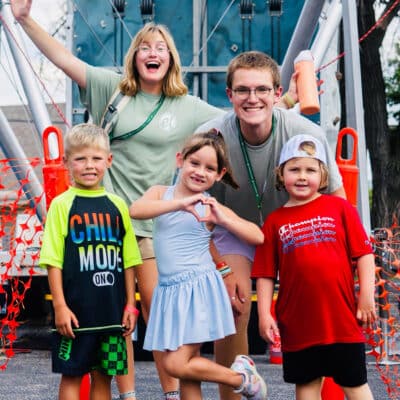 Group of smiling children with two camp counselors posing in front of a climbing structure at an outdoor camp, orange safety net behind them.