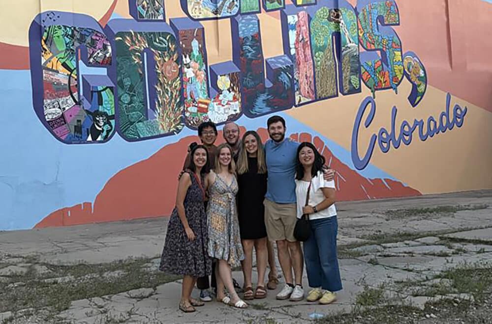 Group of six friends posing in front of a colorful Colorado mural that says 'Colorado' on the wall, wearing casual summer outfits.