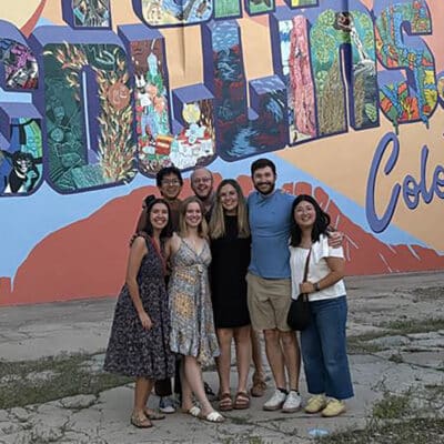 Group of six friends posing in front of a colorful Colorado mural that says 'Colorado' on the wall, wearing casual summer outfits.
