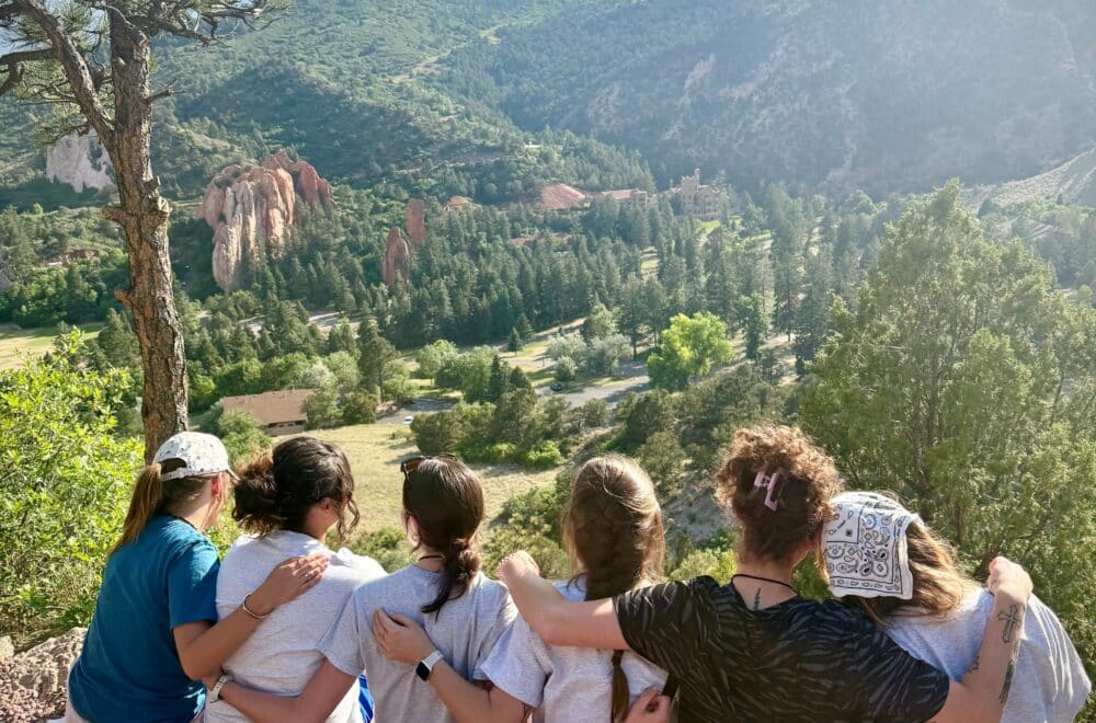 Group of friends sitting with arms around each other on a rocky ledge, facing a scenic valley with pine trees and distant mountains under a blue sky.