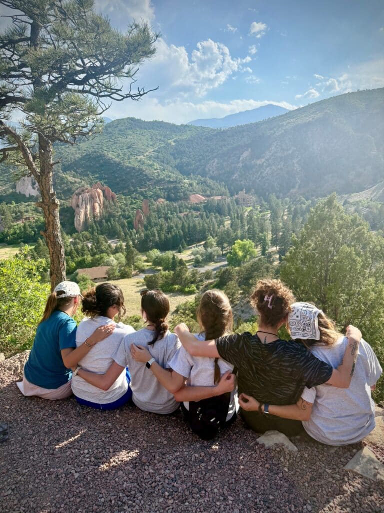 Group of friends sitting with arms around each other on a rocky ledge, facing a scenic valley with pine trees and distant mountains under a blue sky.