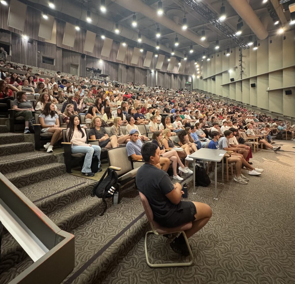 Wide view of a crowded university auditorium filled with students seated in rows, facing the stage.