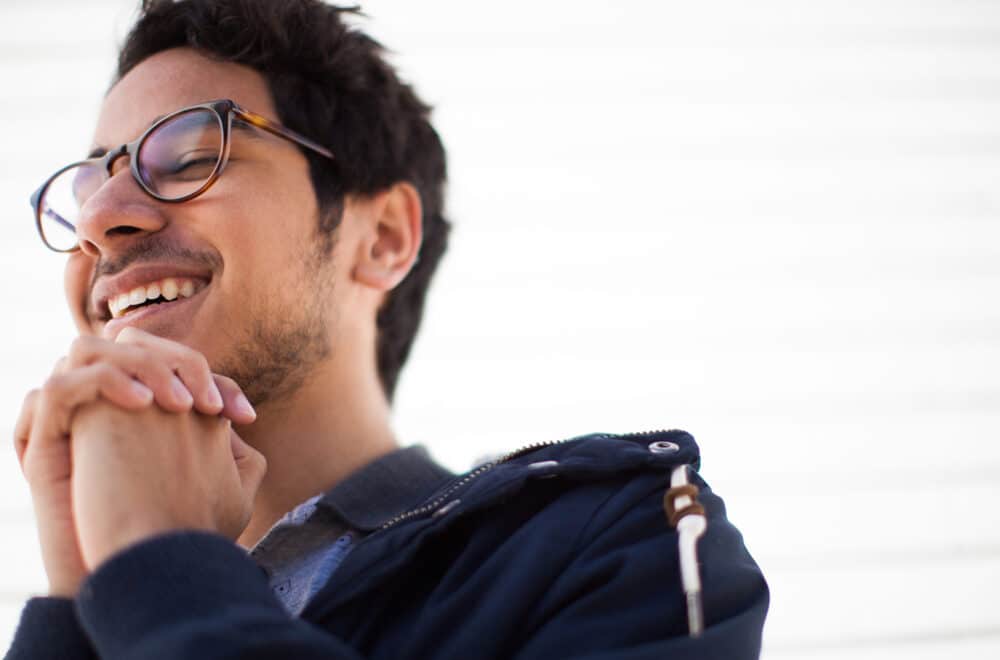 Young man with glasses smiling, hands clasped under his chin, wearing a navy jacket.