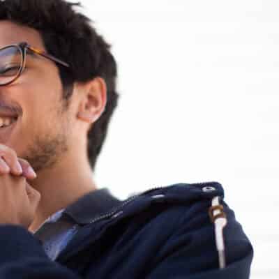 Young man with glasses smiling, hands clasped under his chin, wearing a navy jacket.