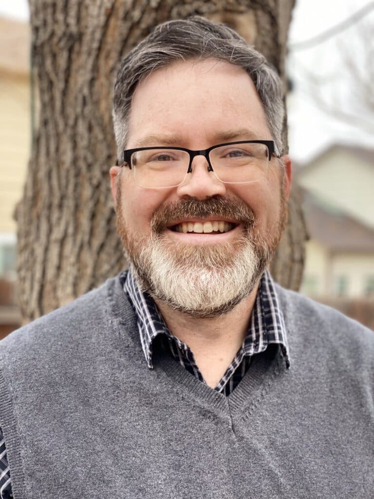 Close-up of a smiling man with glasses and gray beard, outdoors in front of a tree.
