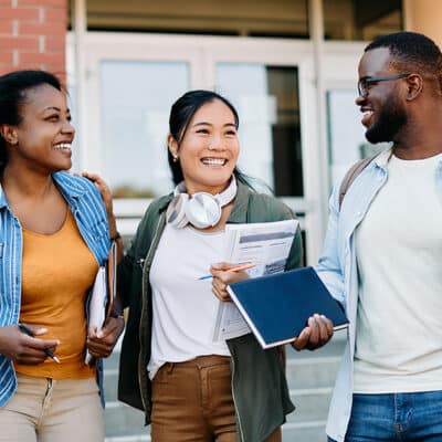 Three diverse college students walking and chatting outside a campus building, carrying notebooks and backpacks.