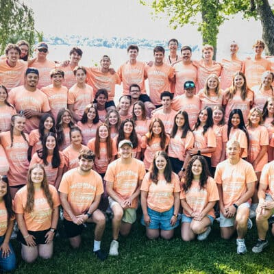 Large group of people wearing peach T-shirts posing outdoors by a lake.