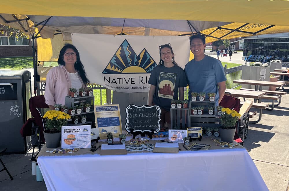 Three volunteers stand at a Native R.I.S.E. booth with potted succulents and informational displays at an outdoor campus event .