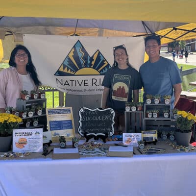 Three volunteers stand at a Native R.I.S.E. booth with potted succulents and informational displays at an outdoor campus event .
