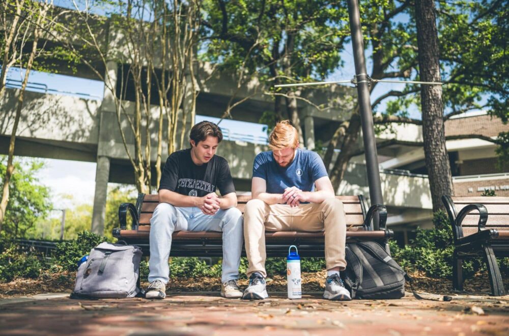 Two college-aged men sit on a bench outdoors on a university campus, with backpacks beside them and a water bottle at their feet.