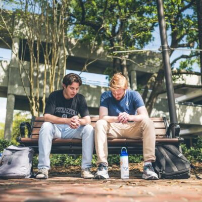Two college-aged men sit on a bench outdoors on a university campus, with backpacks beside them and a water bottle at their feet.