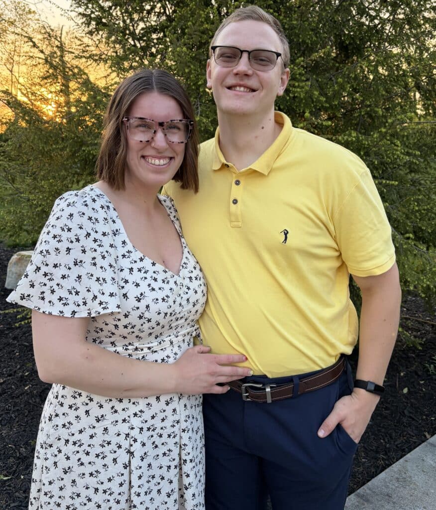 A smiling couple standing close together outdoors at sunset, woman in a patterned dress and man in a yellow polo shirt.