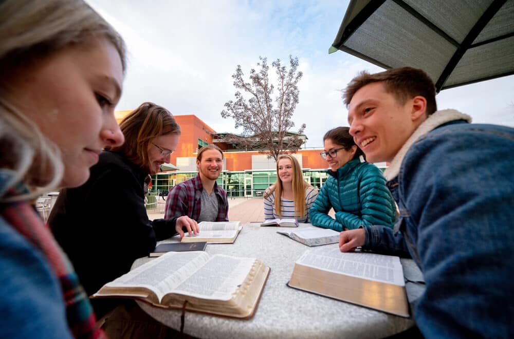 Six students sit around an outdoor round table, studying from open books and notebooks, smiling and talking togetherissuing tags.