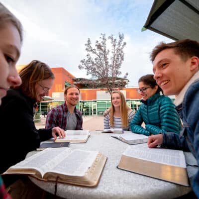 Six students sit around an outdoor round table, studying from open books and notebooks, smiling and talking togetherissuing tags.