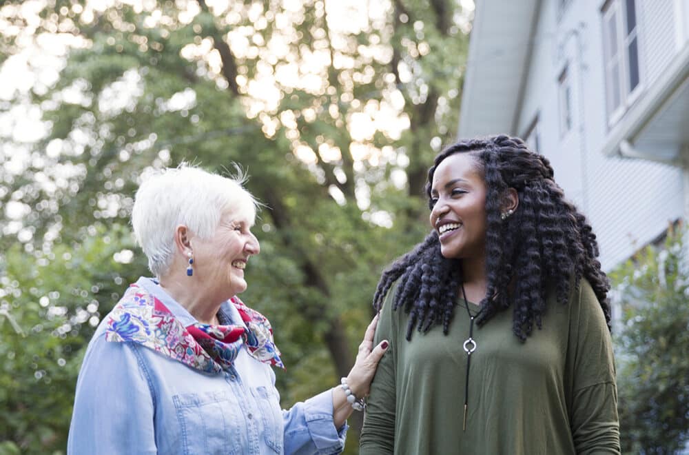 Two women share a warm smile while chatting outside near a light-colored house, one with short white hair in a denim shirt and scarf, the other with long curly hair in an olive green top.