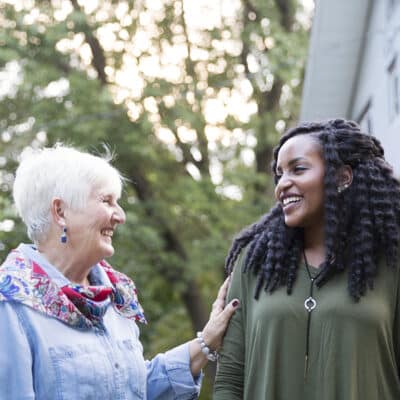 Two women share a warm smile while chatting outside near a light-colored house, one with short white hair in a denim shirt and scarf, the other with long curly hair in an olive green top.
