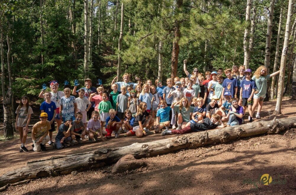 Group of kids of various ages posing together outdoors in a forest, standing and kneeling on fallen logs and dirt path.