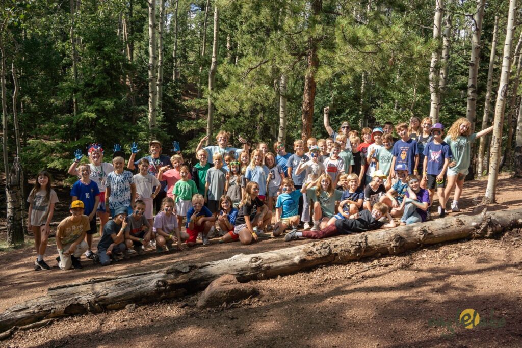 Group of kids of various ages posing together outdoors in a forest, standing and kneeling on fallen logs and dirt path.