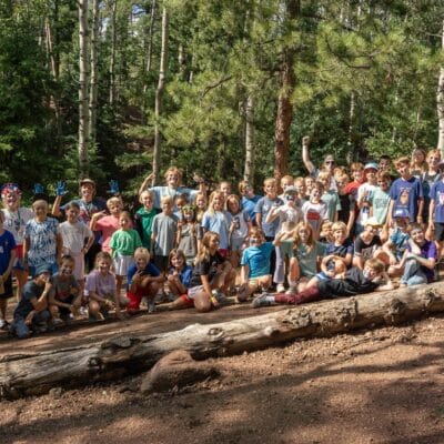 Group of kids of various ages posing together outdoors in a forest, standing and kneeling on fallen logs and dirt path.