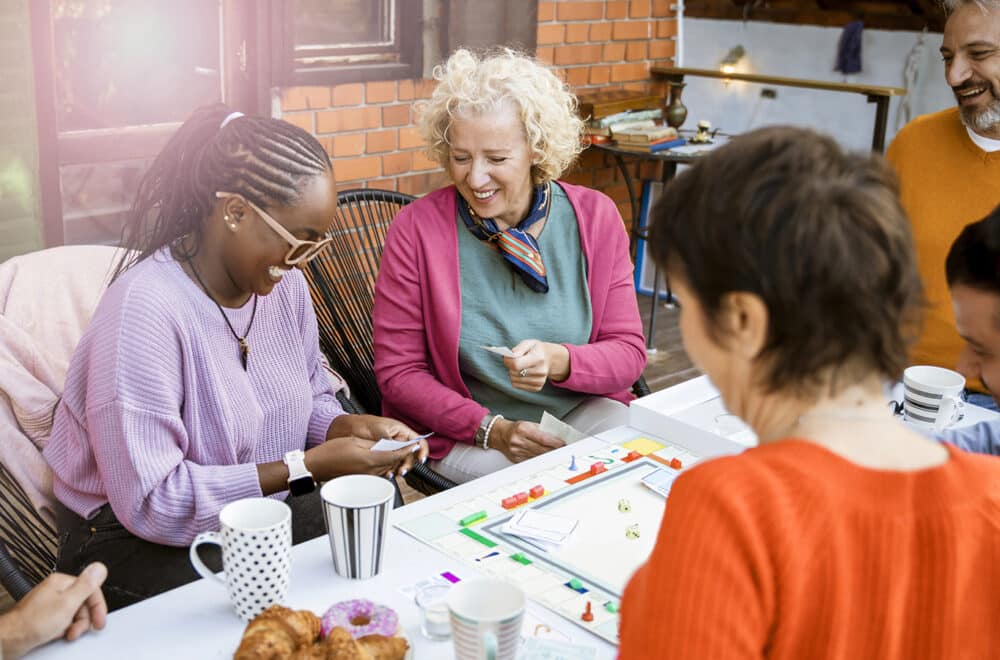 A diverse group of adults gathers around a table to play a board game, smiling and sharing a lighthearted moment.