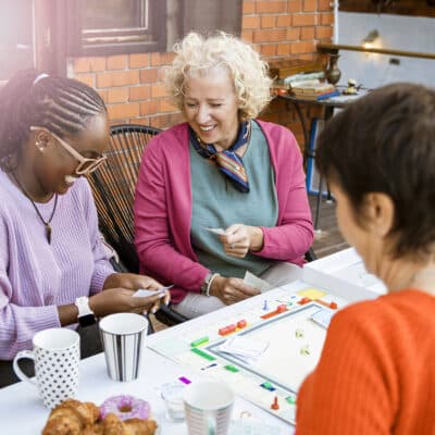 A diverse group of adults gathers around a table to play a board game, smiling and sharing a lighthearted moment.