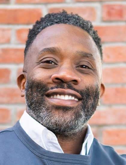 Smiling Black man with short curly hair and gray beard, wearing a white shirt under a blue sweater, brick wall behind.