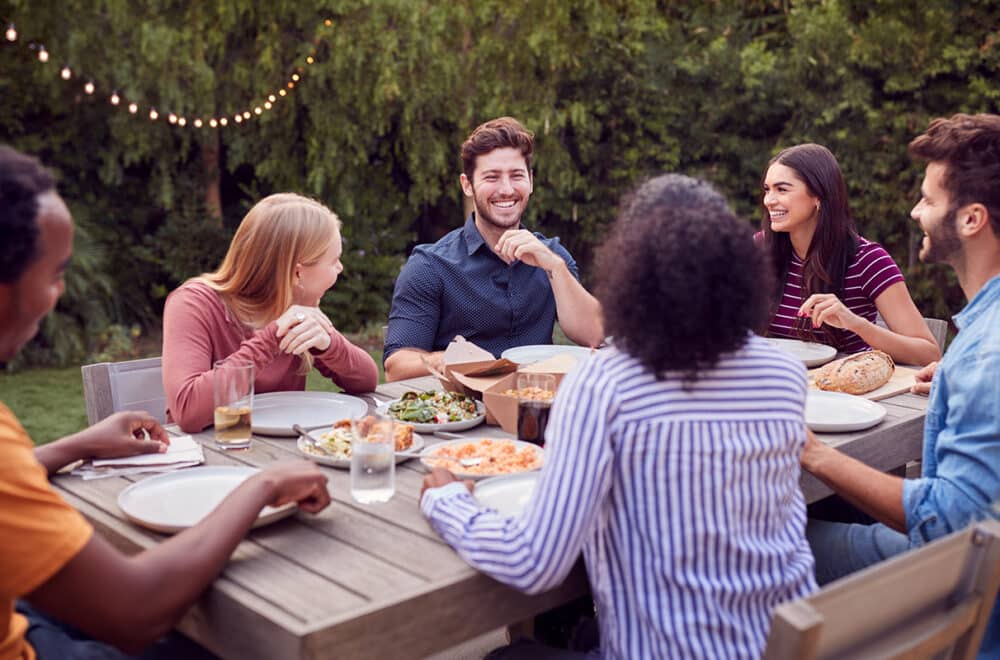 Multi-Cultural Friends At Home Sitting At Table Enjoying Food At Summer Garden Party