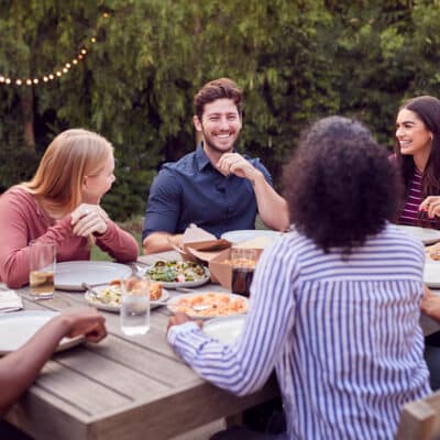 Multi-Cultural Friends At Home Sitting At Table Enjoying Food At Summer Garden Party