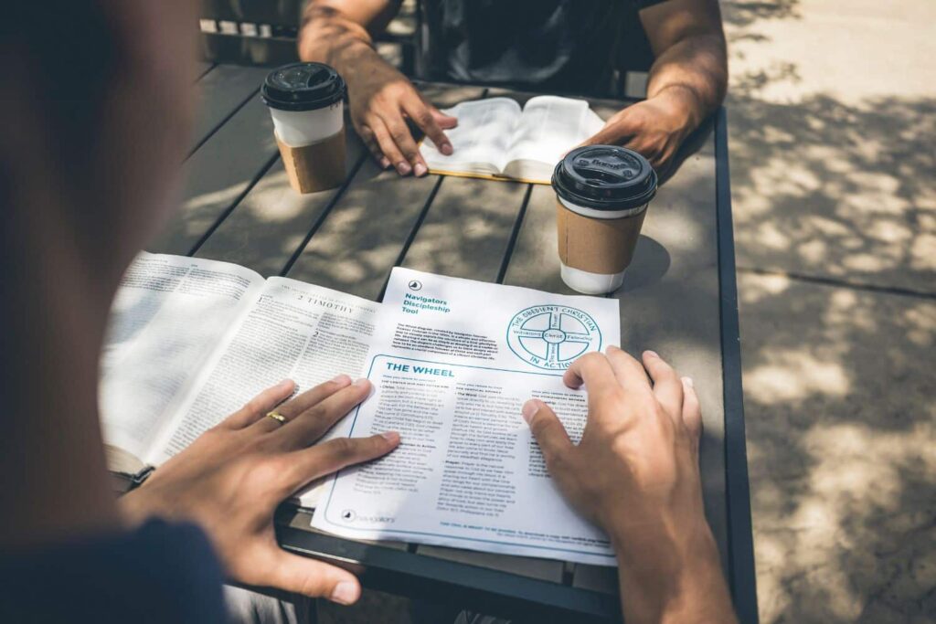 Two people sit at an outdoor table reading a pamphlet; there are two takeaway coffee cups on the table nearby.