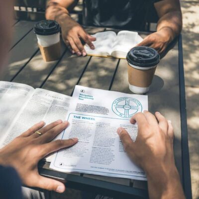 Two people sit at an outdoor table reading a pamphlet; there are two takeaway coffee cups on the table nearby.